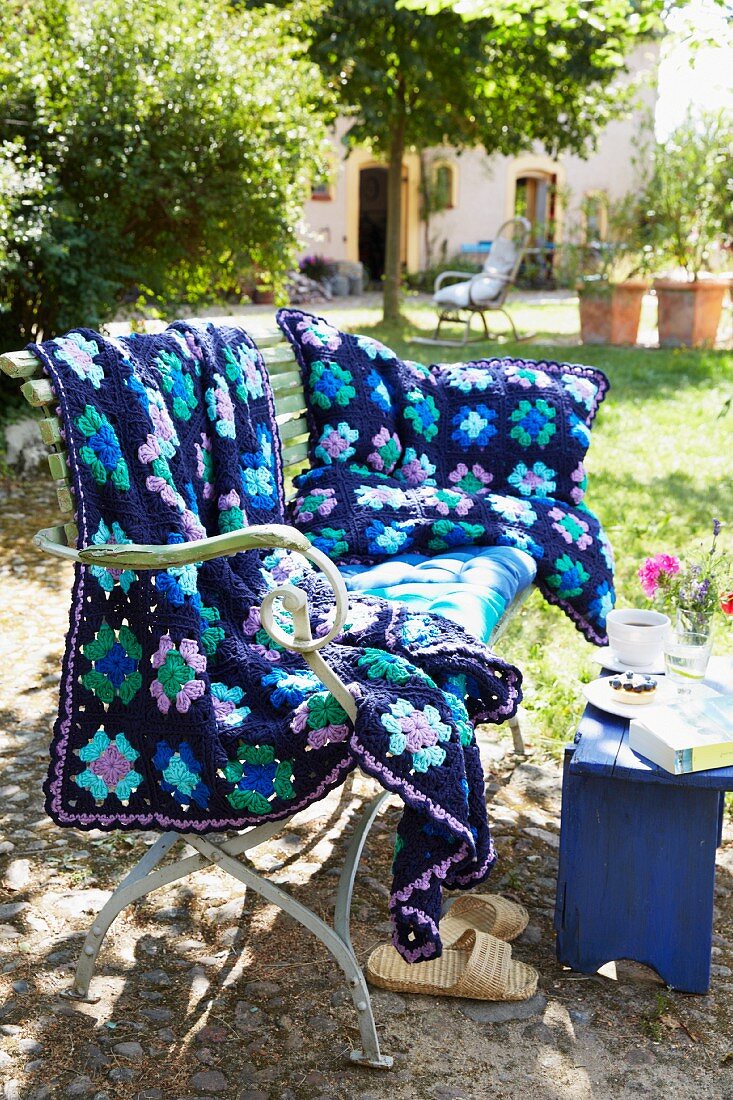 A crocheted blue blanket and cushion cover with a floral pattern on an antique bench in a garden