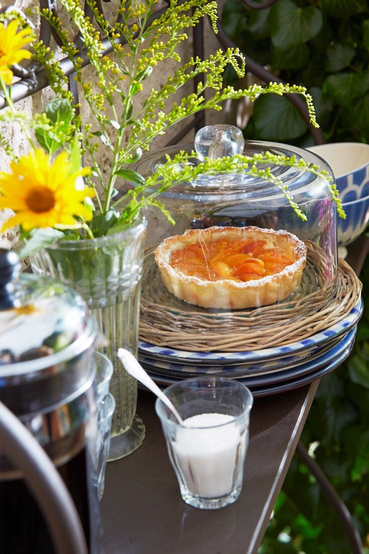 Apricot tart on a wicker plate under a glass cloche with a jug of coffee, French country house crockery and a bunch of meadow flowers on an iron shelf