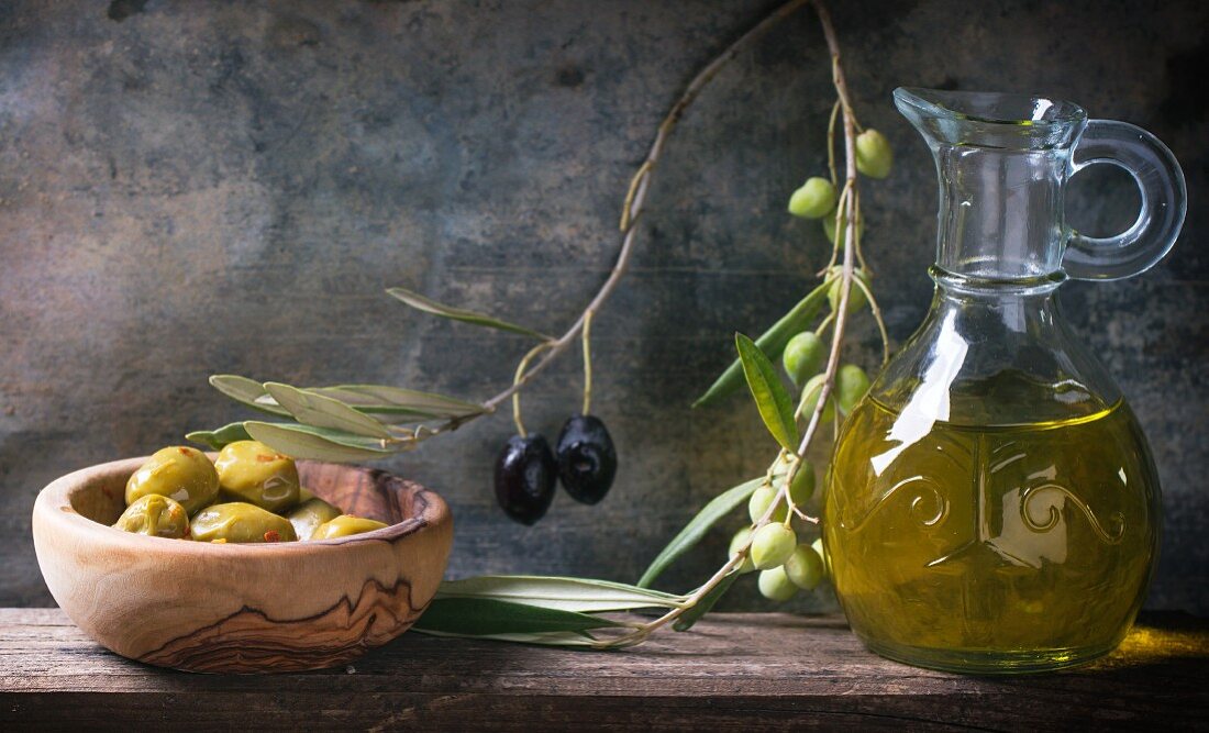 Grüne Oliven in Olivenholzschüssel, Olivenzweige und Karaffe mit Olivenöl