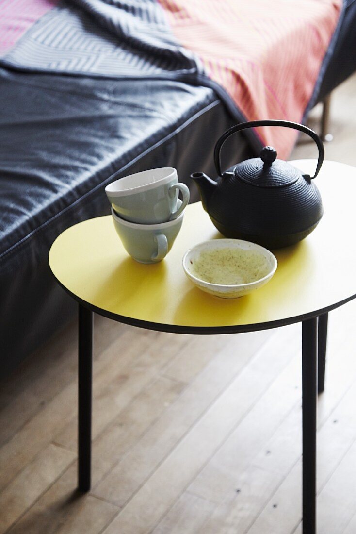 Cast iron teapot and teacups on yellow, kidney-shaped side table in front of black leather sofa