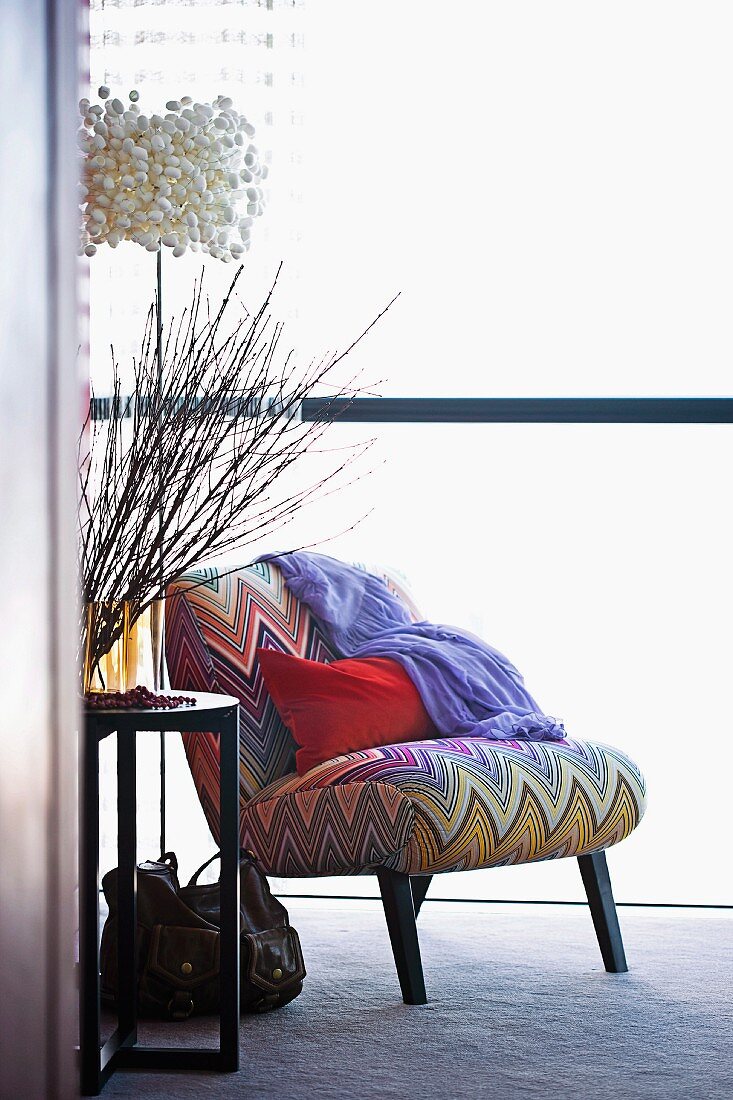 Red cushion on easy chair with zig-zag cover next to vase of twigs on side table and designer lamp
