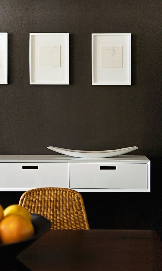 Small drawings in box frames above floating sideboard on dark wall; backrest of wicker chair in foreground