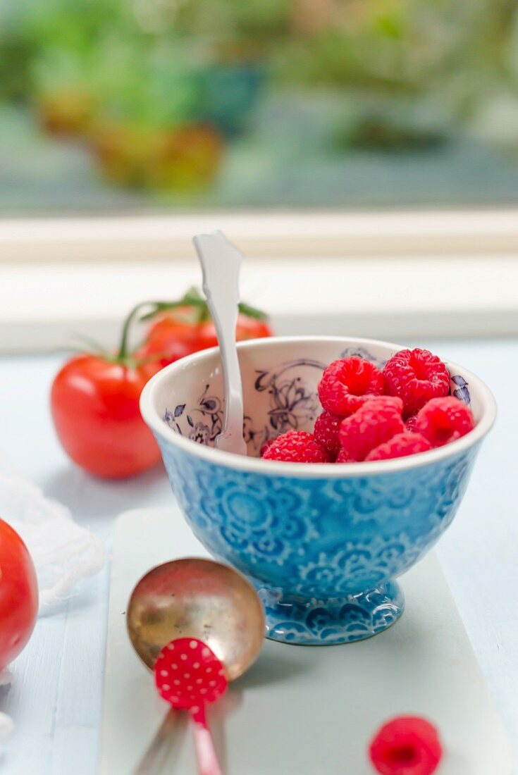 A bowl of fresh raspberries with tomatoes behind it