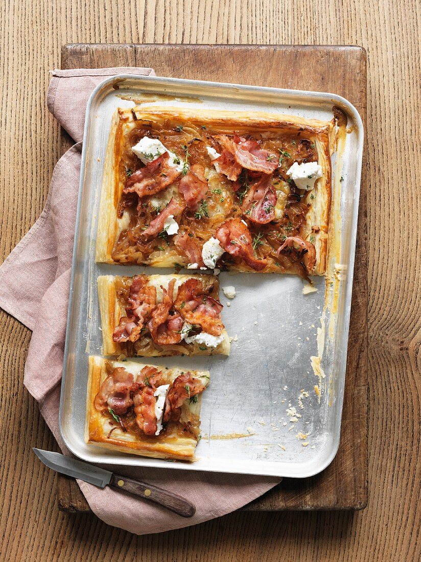 A puff pastry tart on a baking tray with onions, bacon, ricotta and thyme, sliced