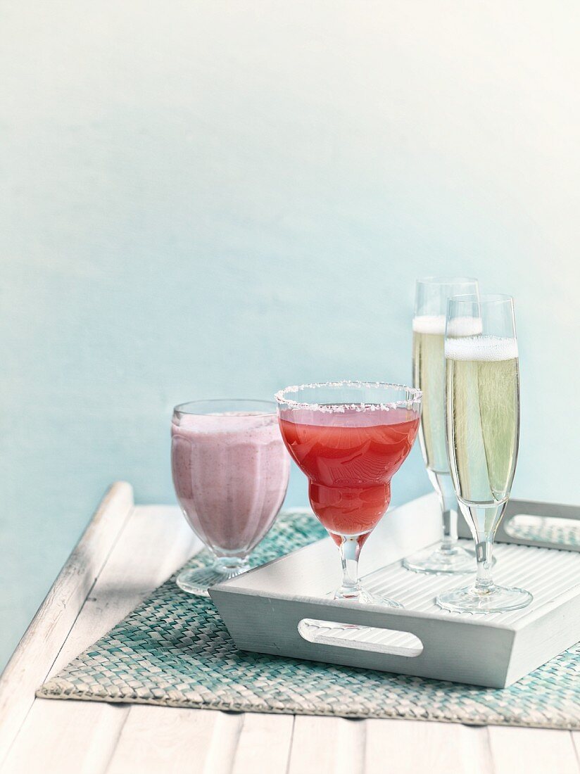 A strawberry milkshake, a strawberry margarita and glasses of champagne on a wooden tray