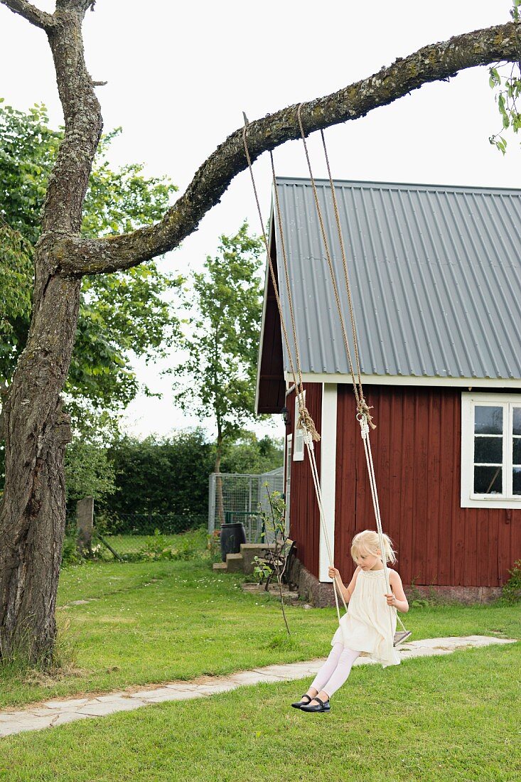 Kleines Mädchen auf Schaukel am Baum, im Hintergrund Holzhaus in sommerlichem Garten