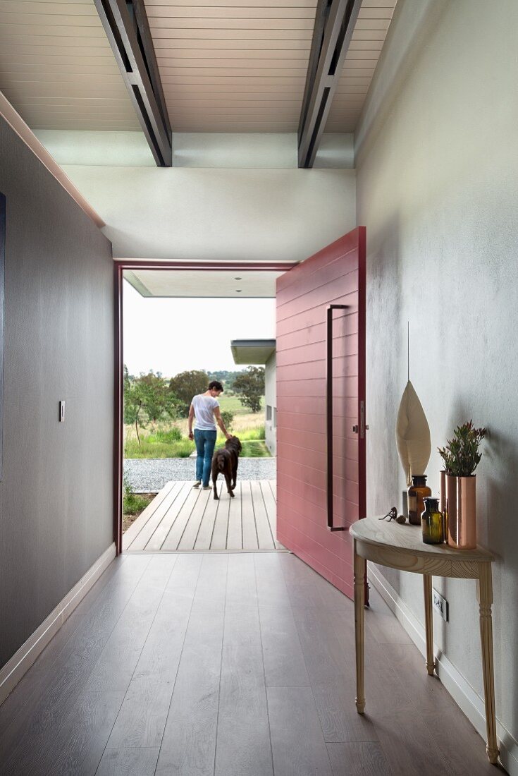 Minimalistischer Eingangsbereich, seitlich Wandtisch, im Hintergrund rot lackierte, offene Haustür und Frau mit Hund