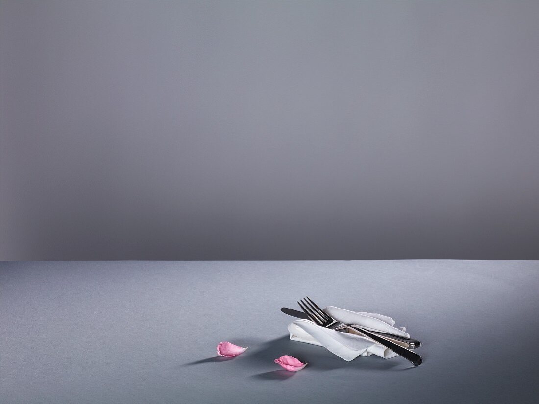 Cutlery on a fabric napkin and pink rose petals on a grey surface