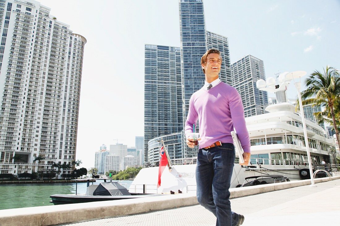 A young business man walking down the street holding a drink and a laptop with a harbour and office blocks in the background