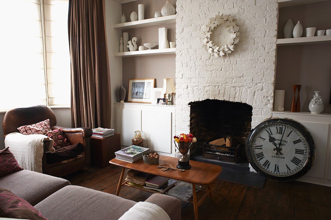 Corner of living room - sofa and armchair around fifties coffee table in front of open fireplace flanked by vases on fitted shelving in niches and antique station clock on floor