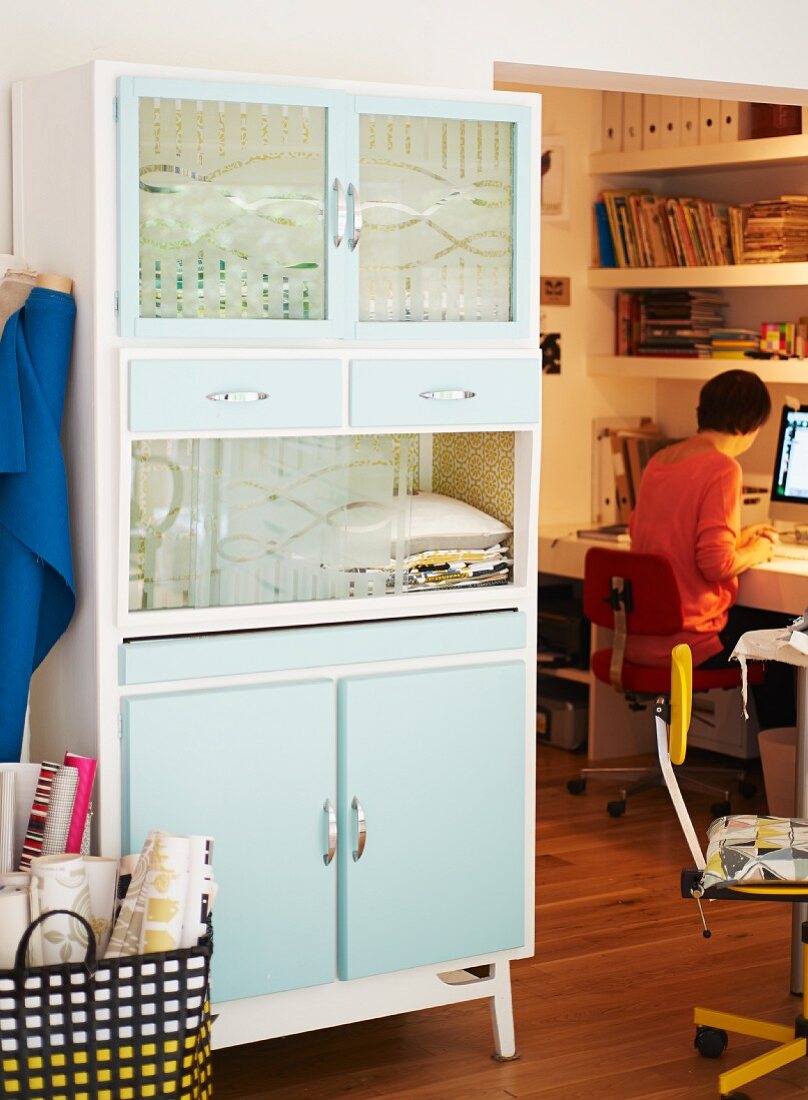 Pastel-coloured fifties cupboard with glass door panels; view of person sitting at desk to one side