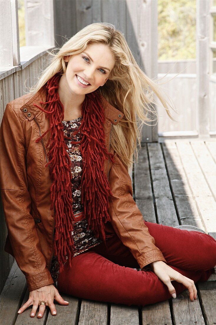 A young blonde woman sitting on a wooden floor wearing a brown leather jacket and a scarf