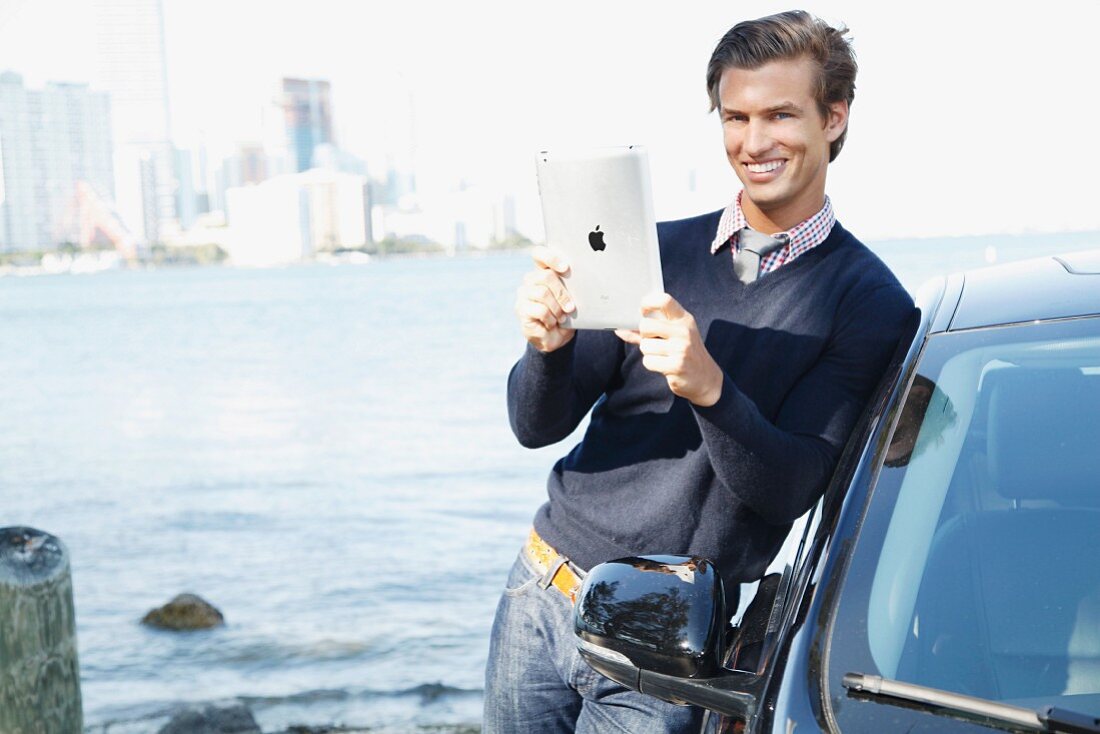 A young man wearing a jumper standing next to a car holding an iPad