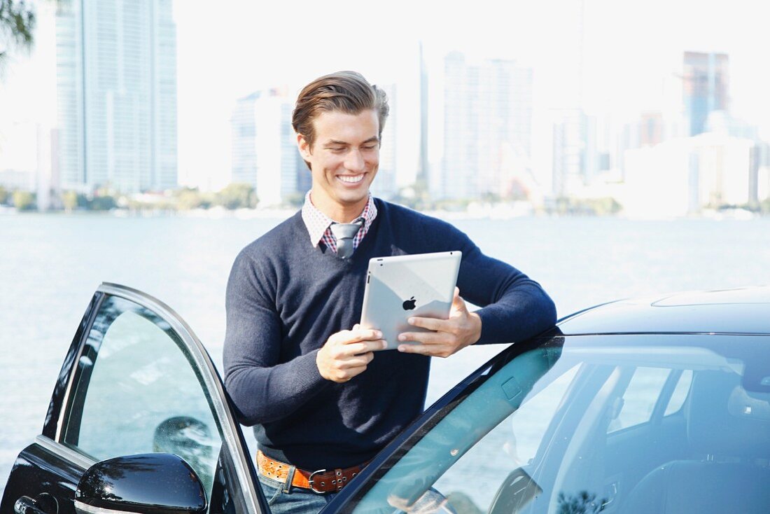 A young man standing next to a car holding an iPad and wearing a jumper