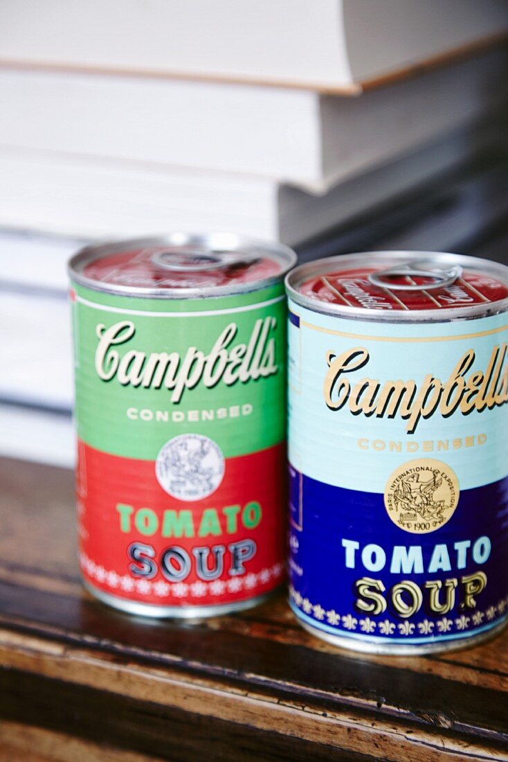 Two tin cans from an Andy Warhol collection in front of stacked books