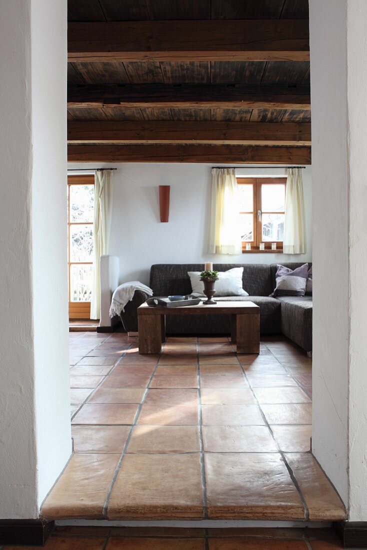 Blick durch offenen Durchgang auf Couchtisch aus Holz und gemütliche Eckcouch in Wohnzimmer eines altes Holzhauses mit Holzdecke