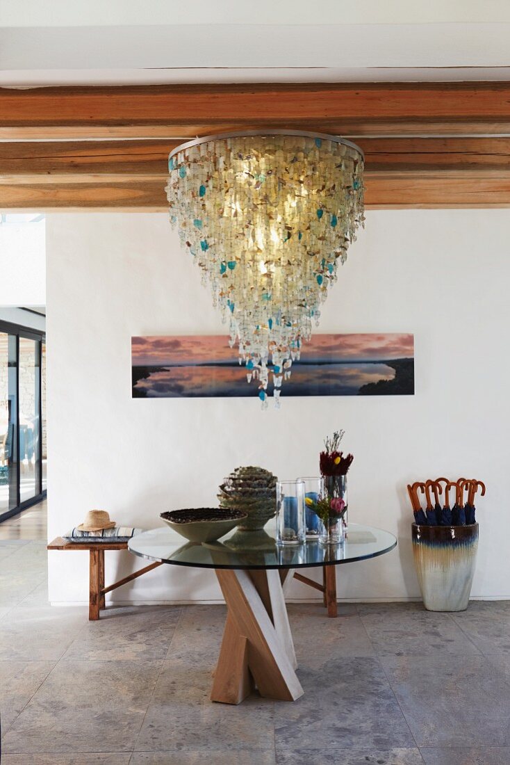 Foyer with opulent chandelier and round glass table top on wooden frame in front of bench and umbrella stand