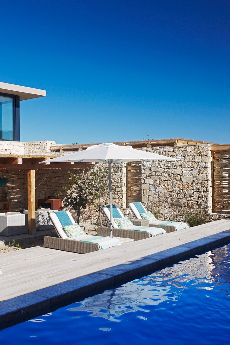 Sonnenliegen und Sonnenschirm auf Holzdeck an blauem Pool vor Natursteinmauer