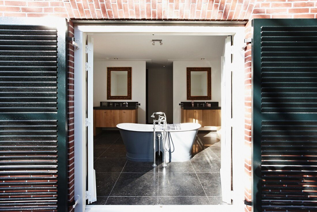 Blick durch offene Tür mit dunklen Türläden auf freistehende Badewanne mit Standarmatur in grossräumigem Bad