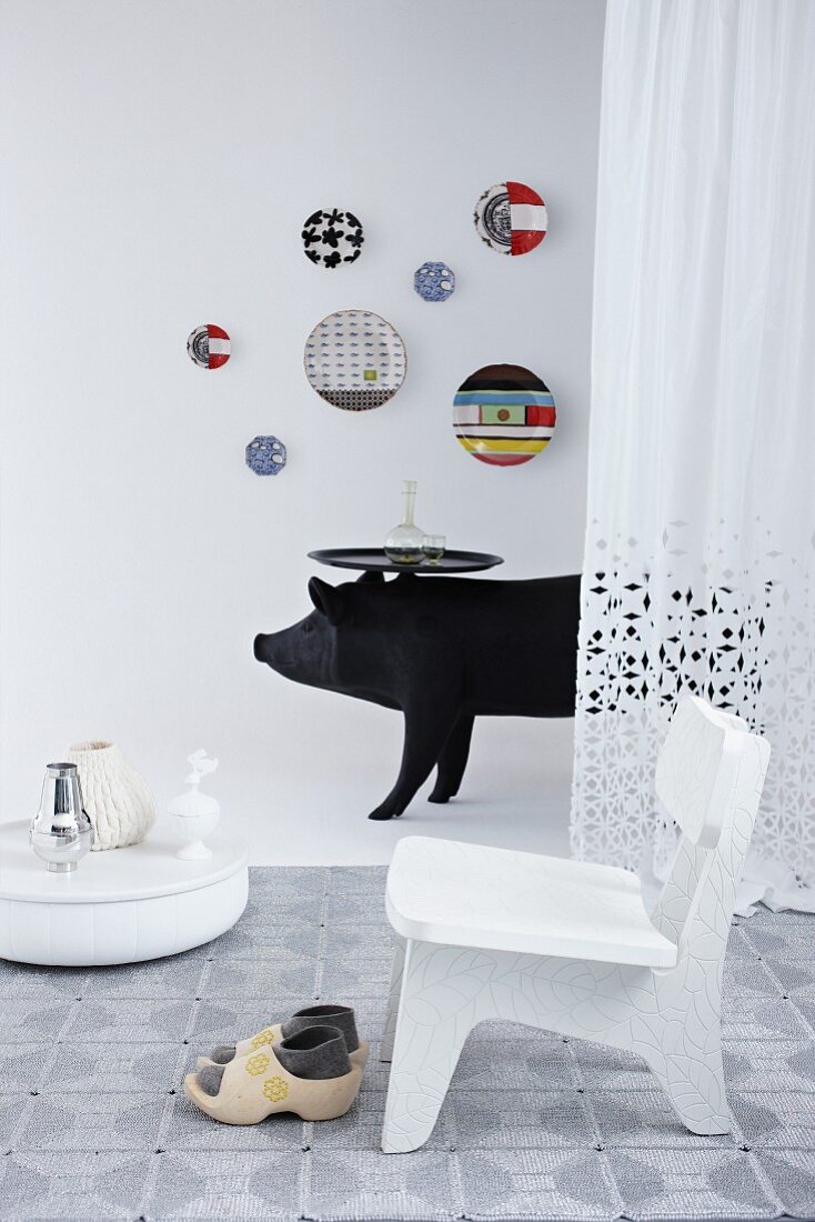 Weisser Stuhl und Holzpantinen auf Teppich, eine schwarze Schwein-Skulptur mit Tabletttisch und bunte Wandteller