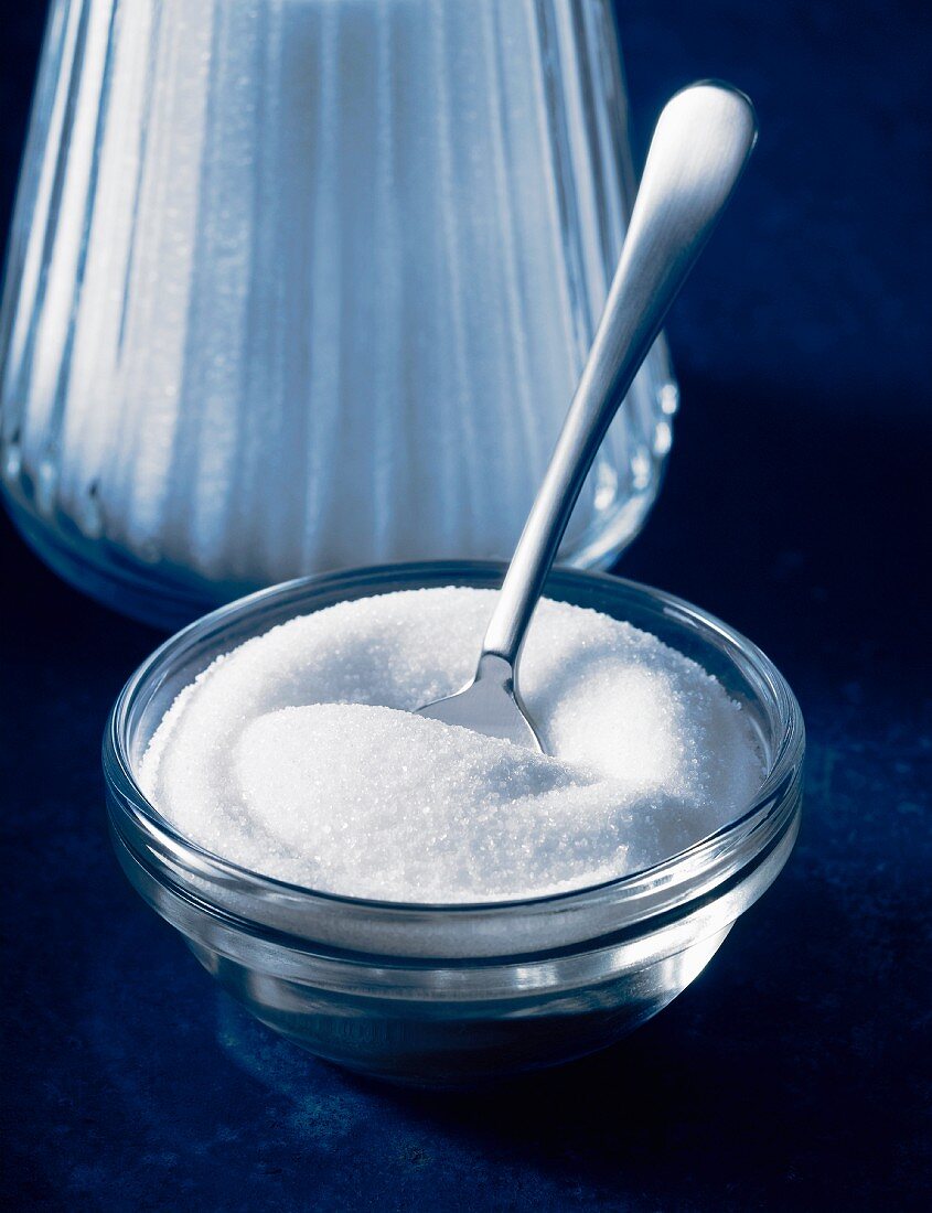 White sugar in a glass bowl with spoon with a sugar shaker behind it