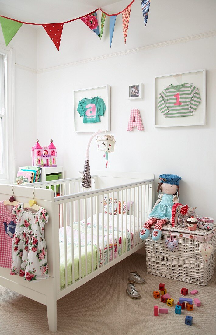 Cot, white wicker trunk, doll and toys in child's bedroom: children's clothing in picture frames on wall