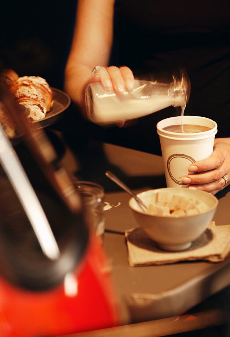 Milk being added to a paper cup of coffee with a bowl of sugar cubes in the foreground