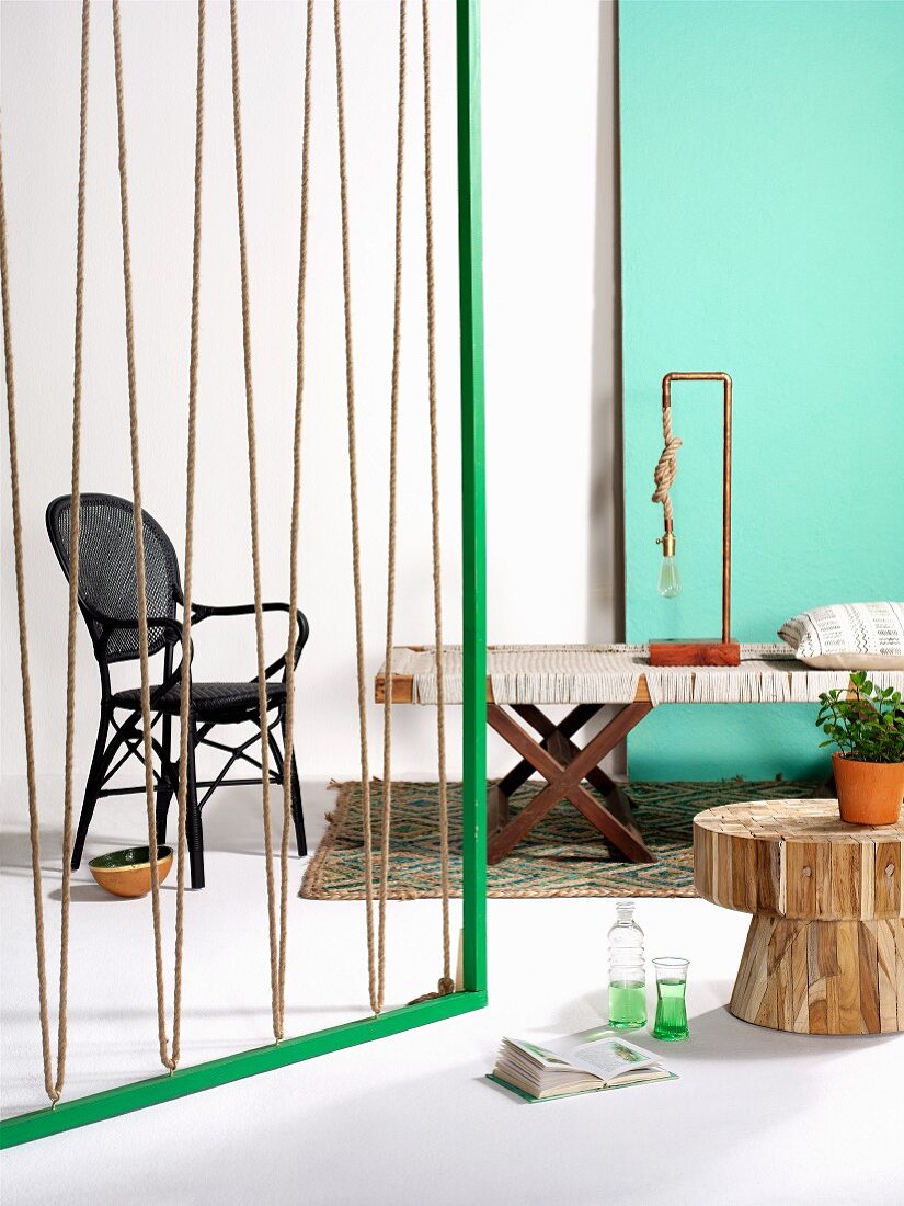 Green wooden frame strung with rope as partition, bench with fabric seat, lamp made from copper piping and rope