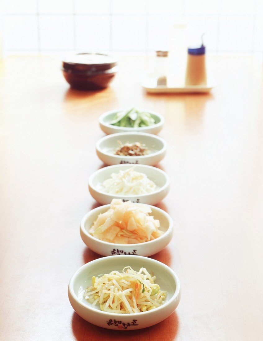 Five Korean side dishes in white bowls with soy sauce, salt and chilli powder in the background