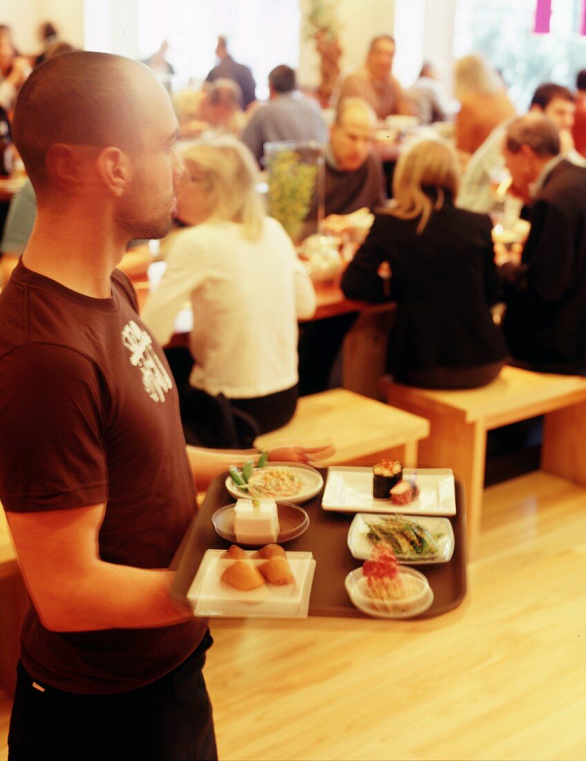 A waiter carrying a tray of various dishes through a crowded Korean restaurant