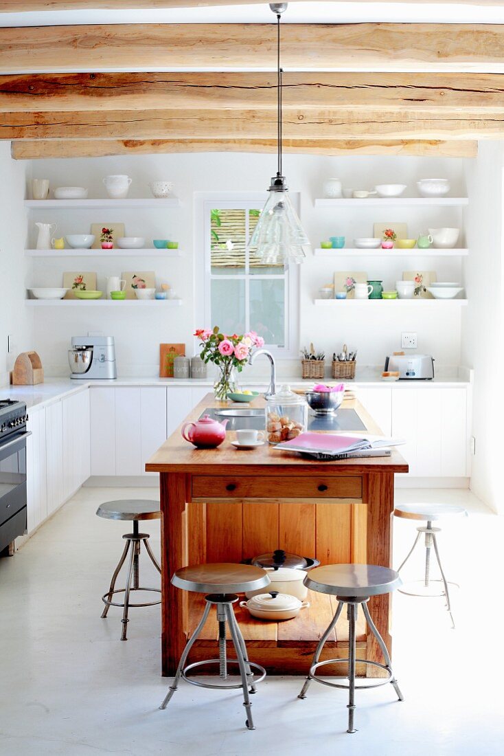 Vintage metal stools around free-standing, solid-wood kitchen island under wood-beamed ceiling with modern fitted shelves and cupboards in background
