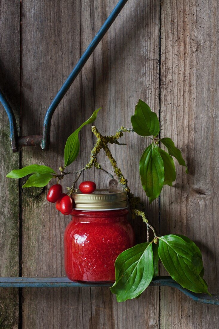 A jar of home-made cornelian cherry jam with a sprig against a wooden wall