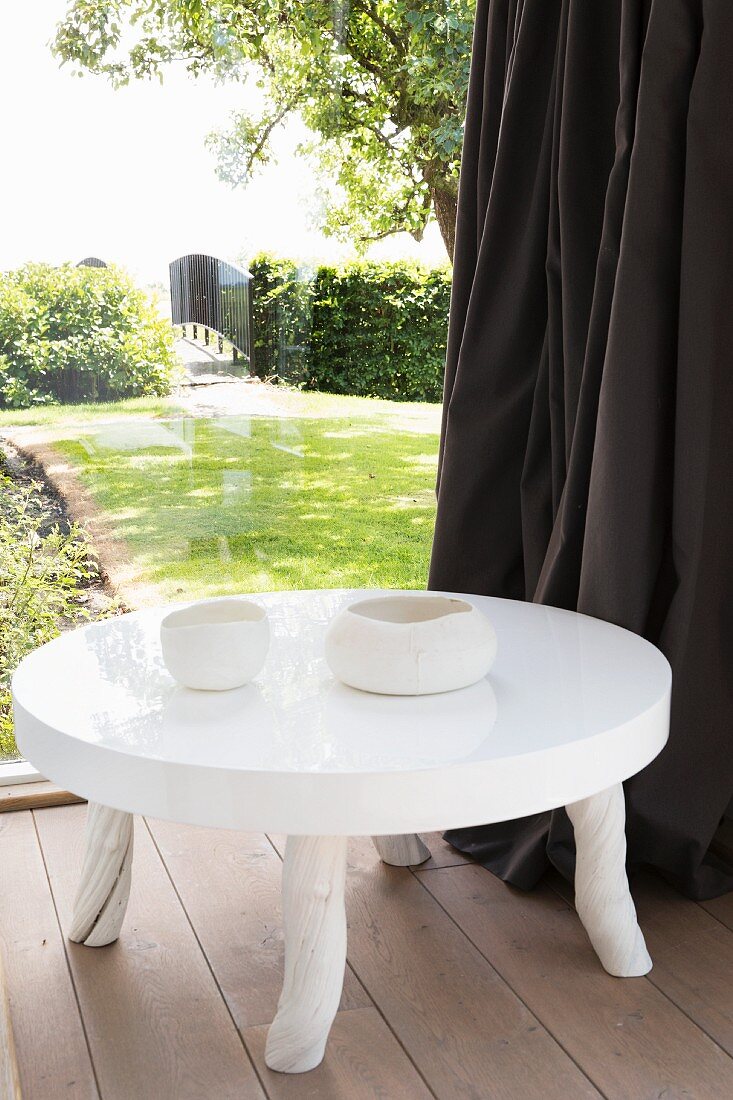 Low, white table with glossy top and rustic legs next to glass wall with view into summery garden