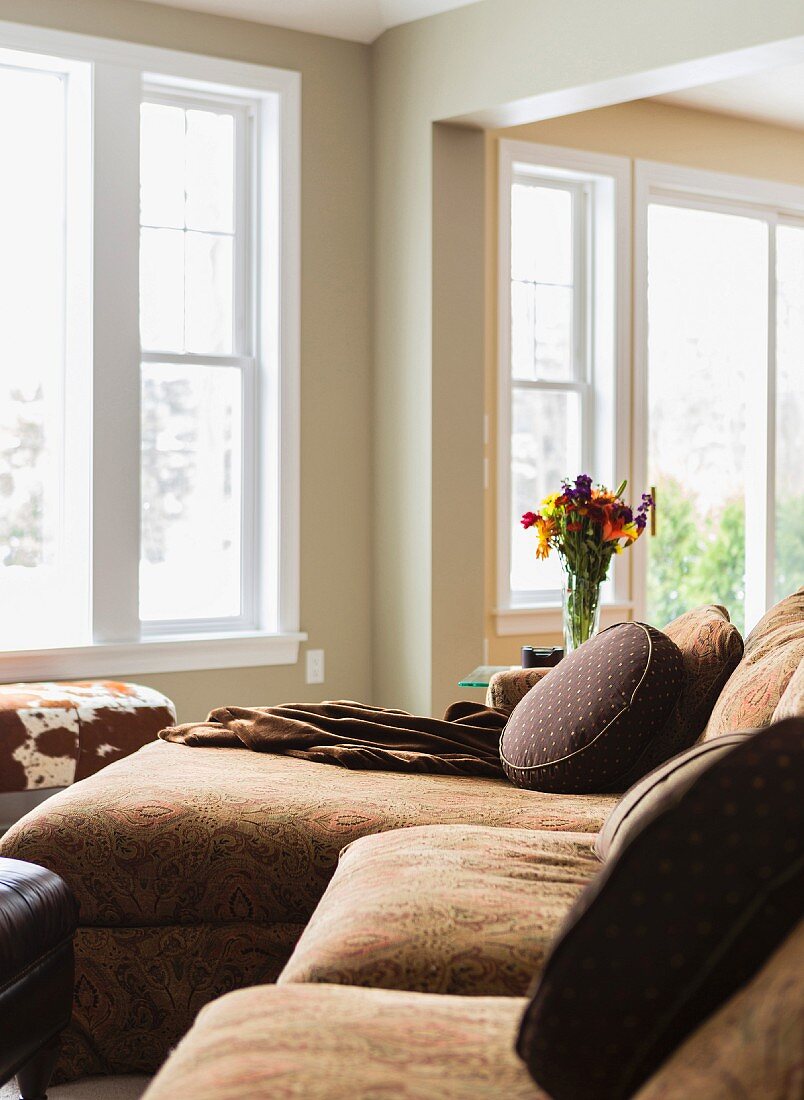 Grosse Couch mit runden, dunkelbraunen Zierkissen; davor ein Polstertisch und daneben ein Strauss bunter Blumen auf einem Beistelltisch