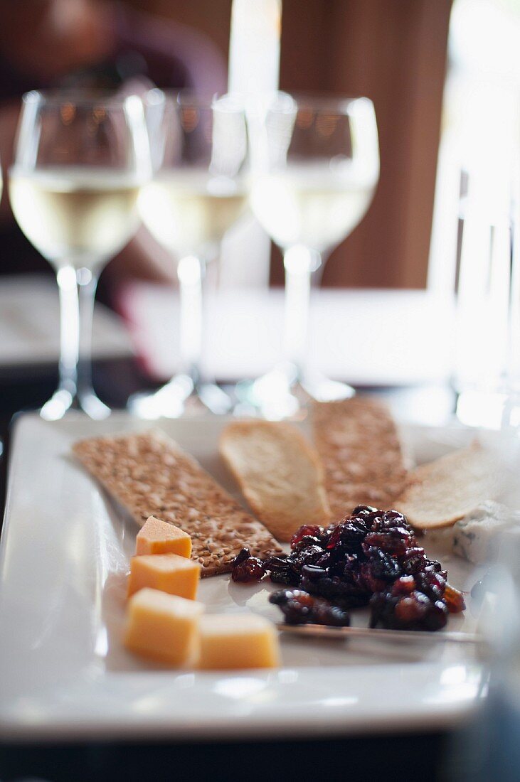 An appetizer platter featuring cheese, crisp breads and raisins with glasses of white wine in the background