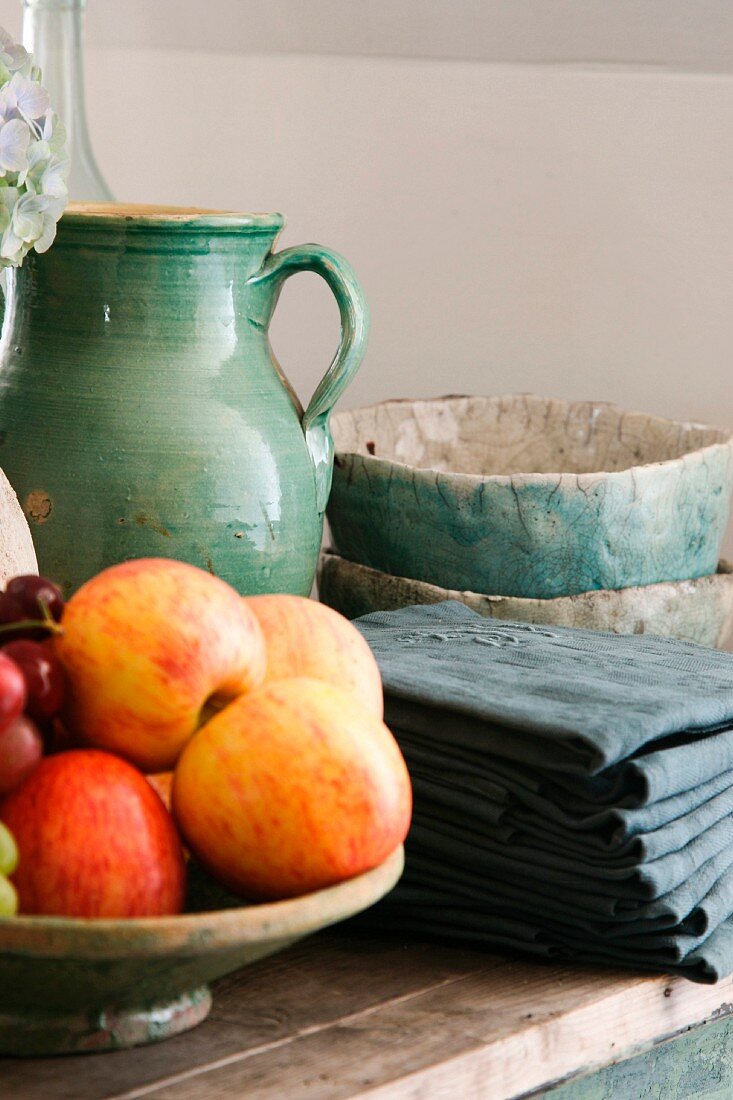 Bowl of apples next to folded dark grey cloths and ceramic pots