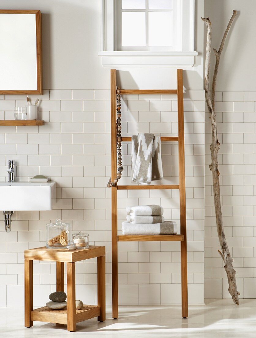 Hocker aus Teakholz neben leiterartigem Handtuchhalter an weisser Fliesenwand, seitlich getrockneter Ast im Badezimmer