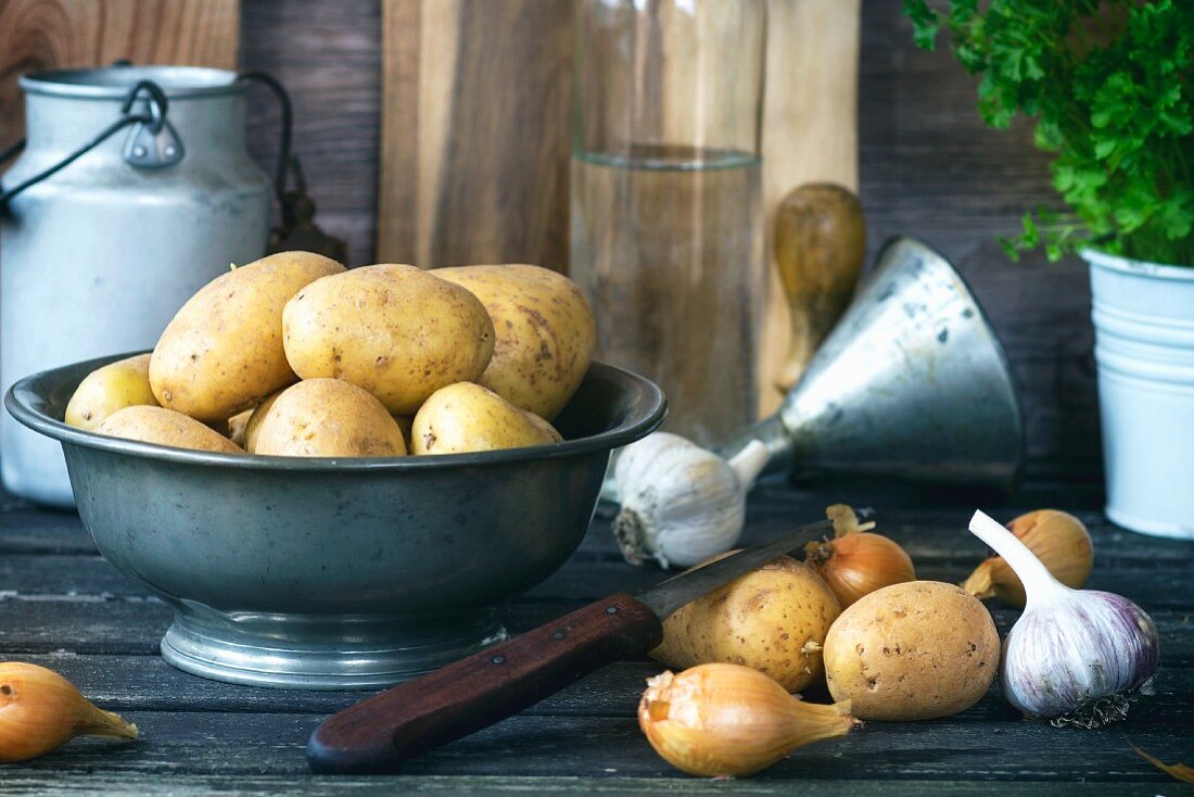 Potatoes in a metal bowl next to onions and garlic on an old wooden table
