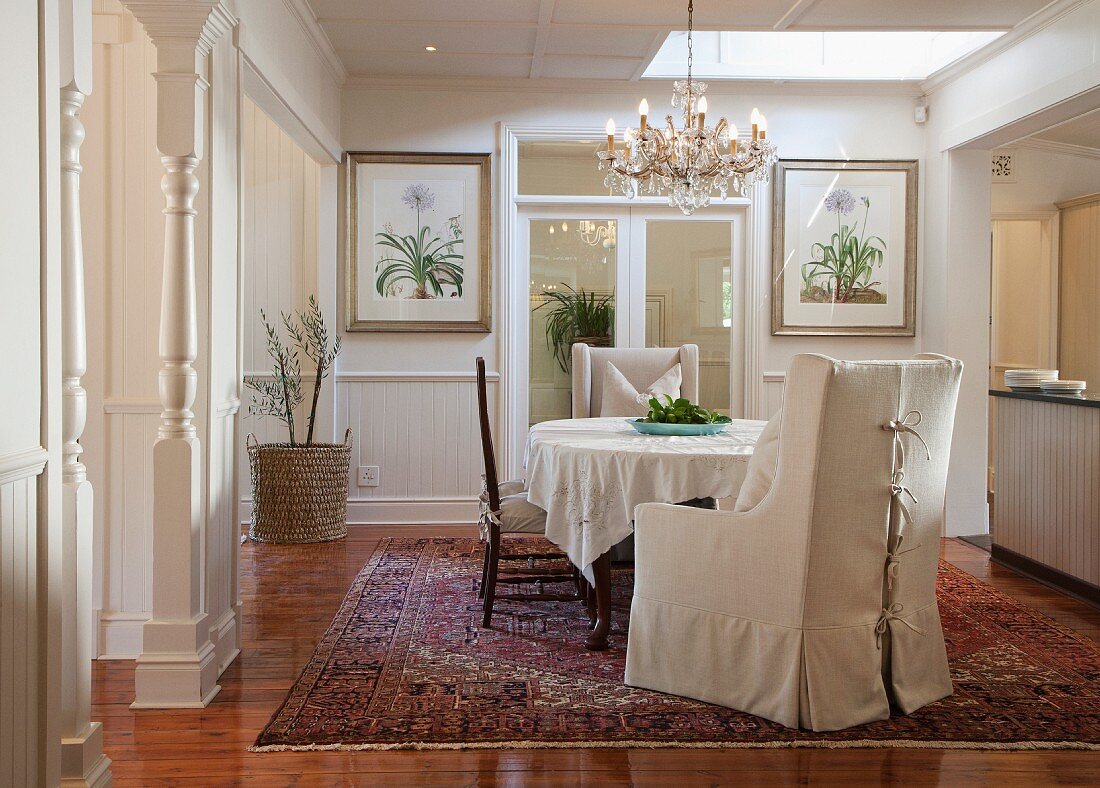 Armchair with pale loose cover, chairs around table with tablecloth and chandelier in traditional, elegant dining room