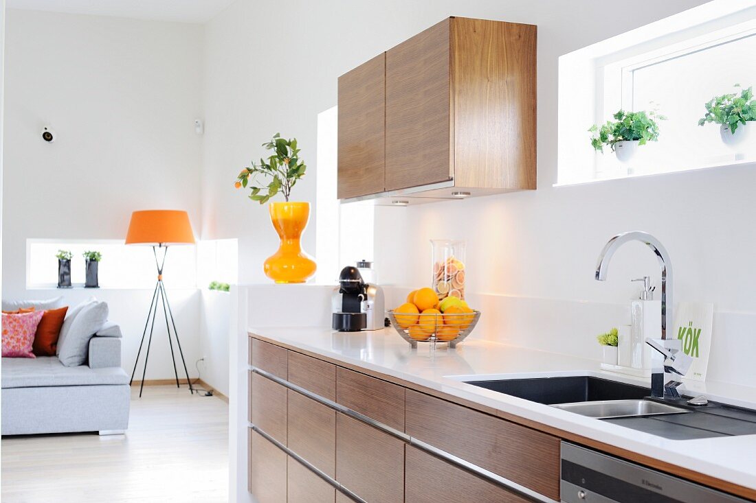 Open-plan kitchen with wooden cabinets and white worksurface; standard lamp with orange lampshade in lounge area in background