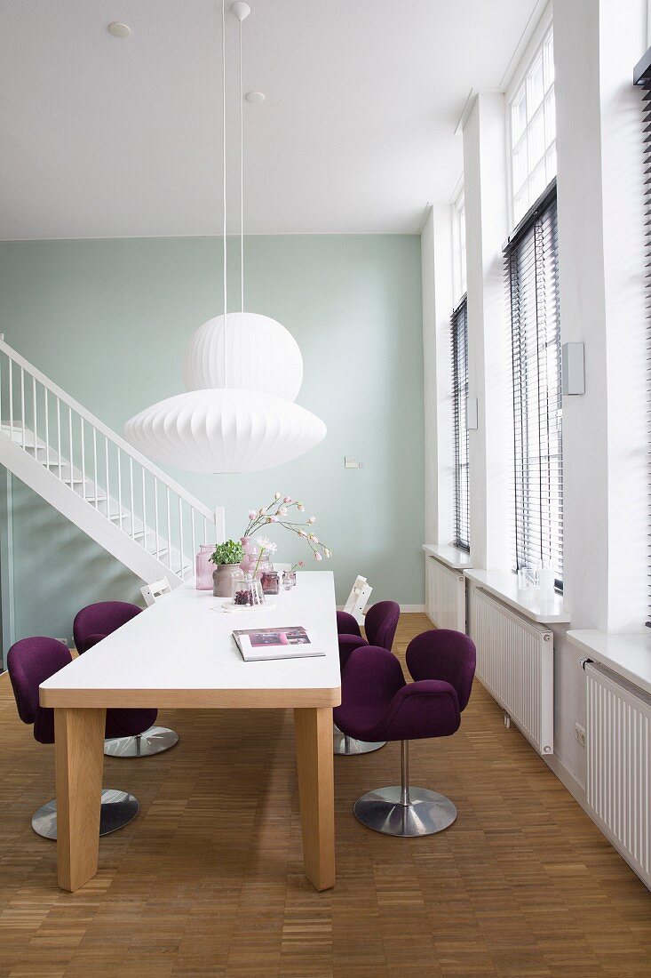 Purple, designer swivel chairs at solid wooden table below white pendant lamps; staircase in background