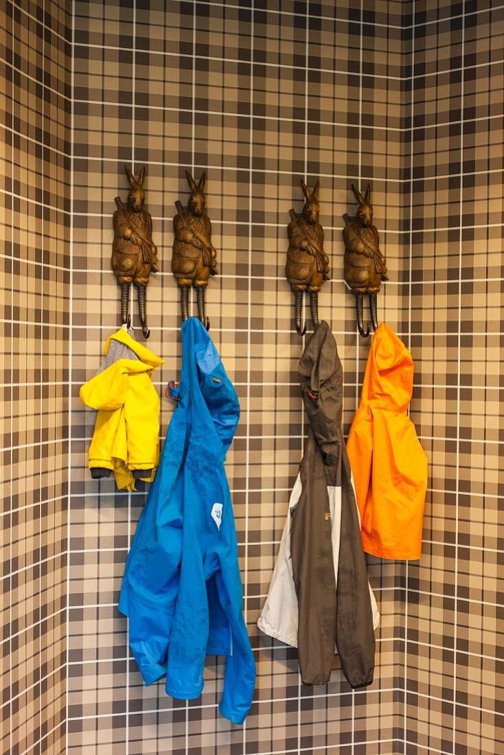 Farbige Regenjacken, aufgehängt an Kleiderhaken in Form von menschenähnlichen Hasenfiguren vor braun kariertem Hintergrund