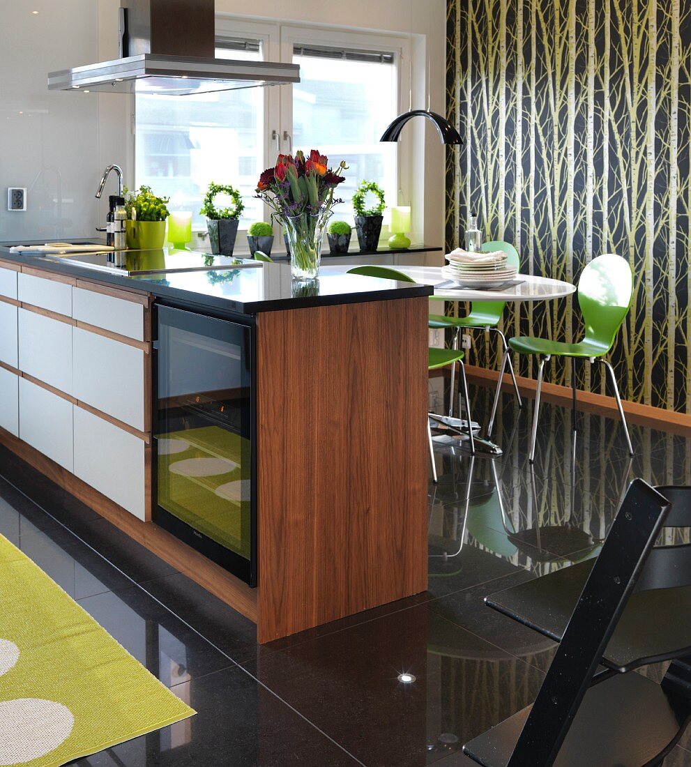 Walnut island counter in front of dining area with green chairs on glossy black-tiled floor and tree-patterned wallpaper