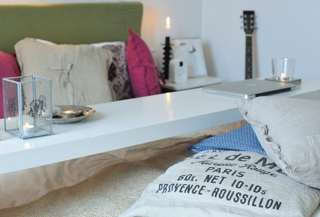 Laptop and lit candles on narrow over-bed table above bed with many scatter cushions