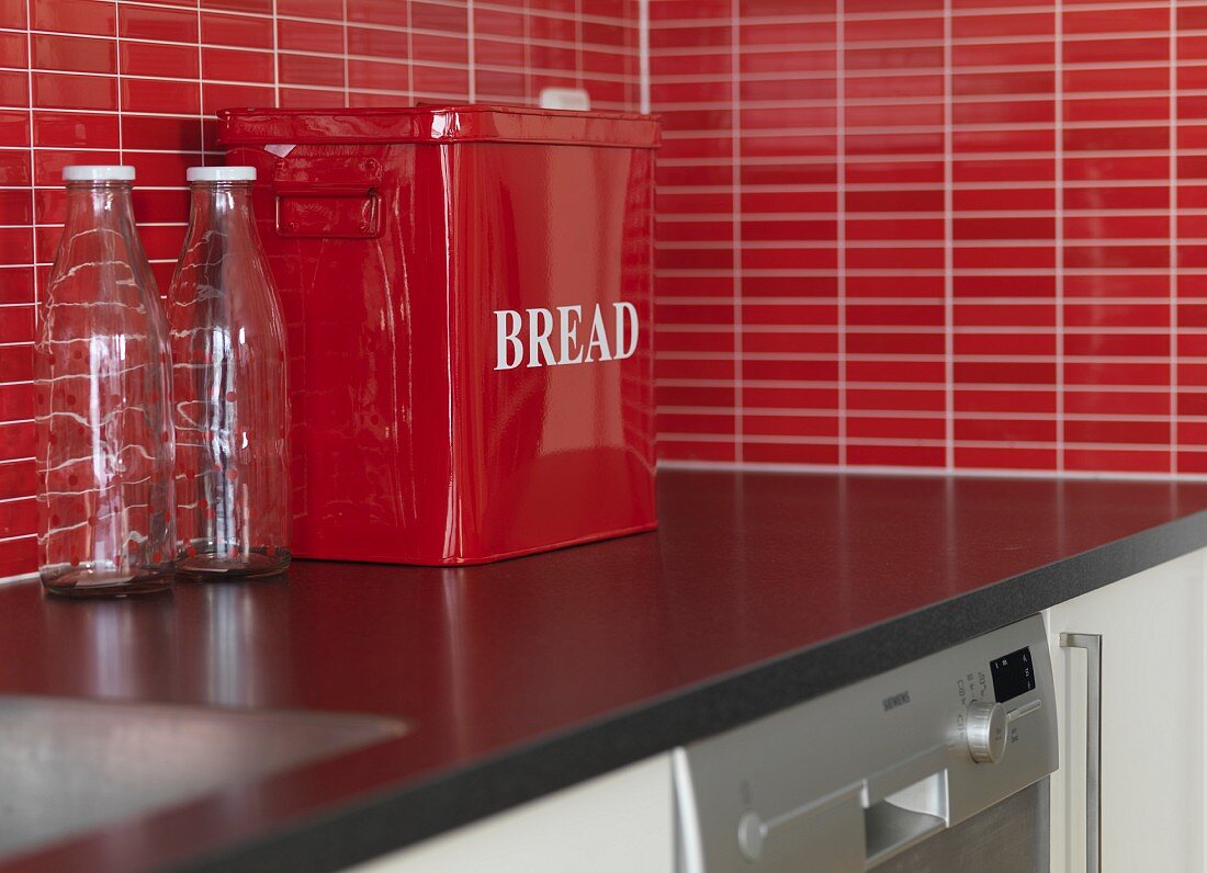 Empty milk bottles and red metal bread bin on kitchen worksurface with splashback of red mosaic tiles