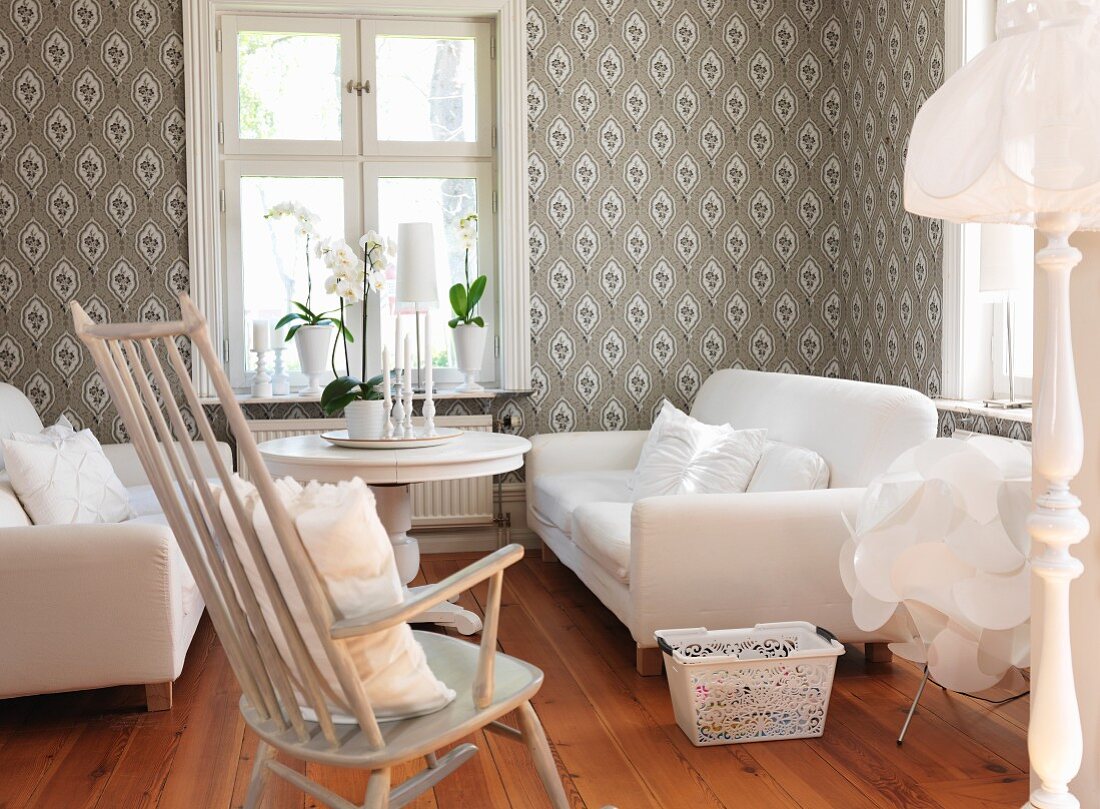Gemütliches Wohnzimmer - weiße Sofagarnitur und Schaukelstuhl um Tisch am Fenster, an Wand gemusterte Tapete