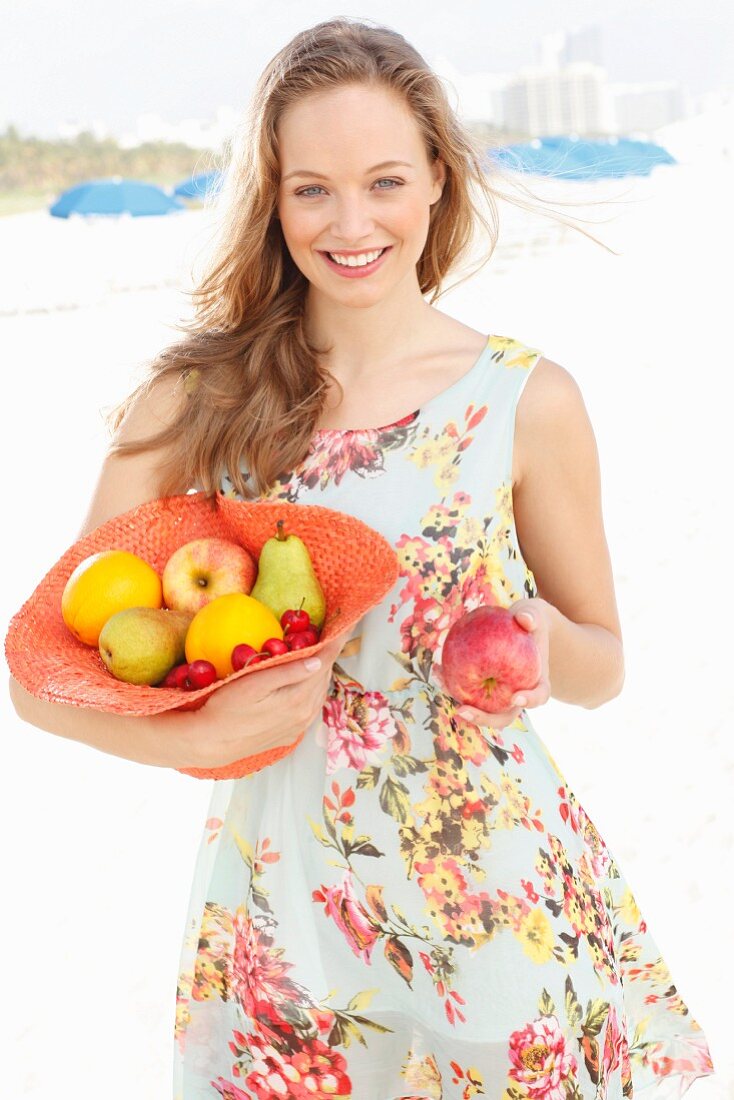 Junge Frau im Sommerkleid hält mit Obst gefüllten Hut und bietet einen Apfel an