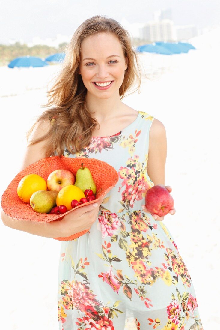 A young woman wearing a summer dress holding a hat filled with fruit and holding out an apple