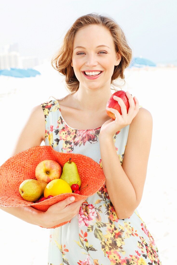 Junge Frau im Sommerkleid hält mit Obst gefüllten Hut und einen Apfel
