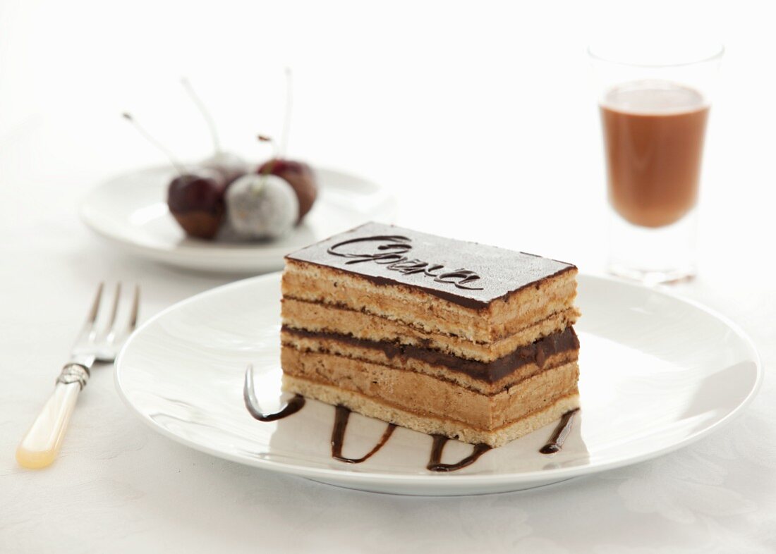 A slice of Opera Cake with cherries and a glass of chocolate liqueur in the background